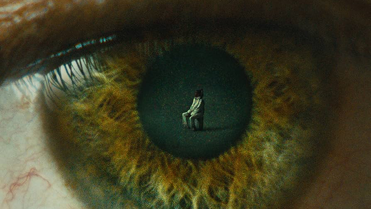 Détail de la couverture du vinyle "L'Enfer" de Stromae. Gros plan d’un œil humain aux teintes vertes et dorées. Au centre de la pupille, une petite silhouette humaine est visible, assise seule sur une chaise dans un espace sombre, créant un effet surréaliste