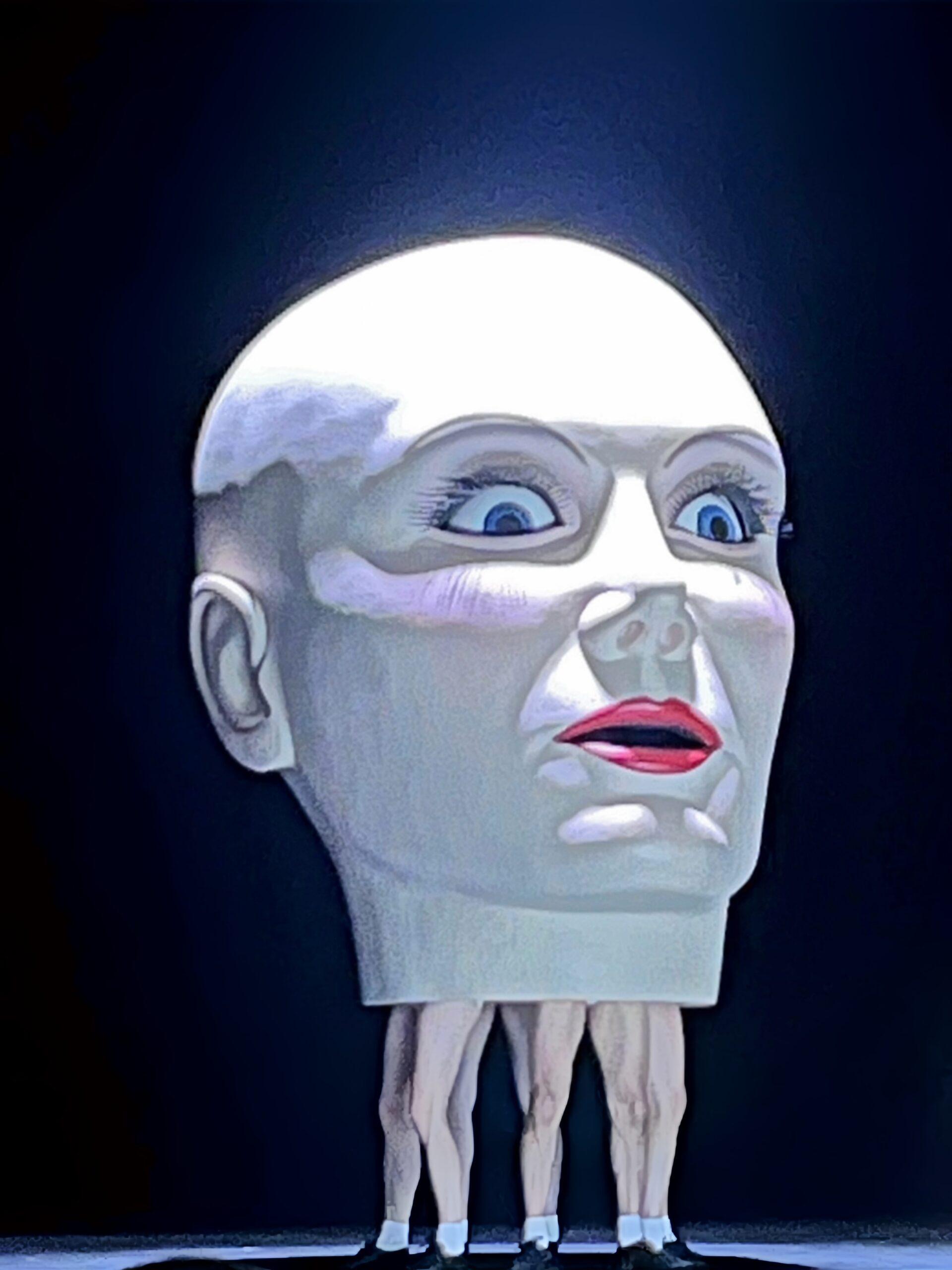 Photo prise lors de la représentation de "La Femme sans ombre" de Richard Strauss au Festival d’Aix-en-Provence en 2026. Une structure-sculpture représentant une énorme tête humaine féminine blanche, chauve, aux yeux bleus grands ouverts et à l'expression figée, est portée par quatre danseurs. Seules les jambes des danseurs sont visibles. Elles sont nues chaussées de chaussettes blanches et des chaussures noires. La scène est dans la pénombre, la tête se dégage parfaitement, l’atmosphère est inquiétante et étrange.
