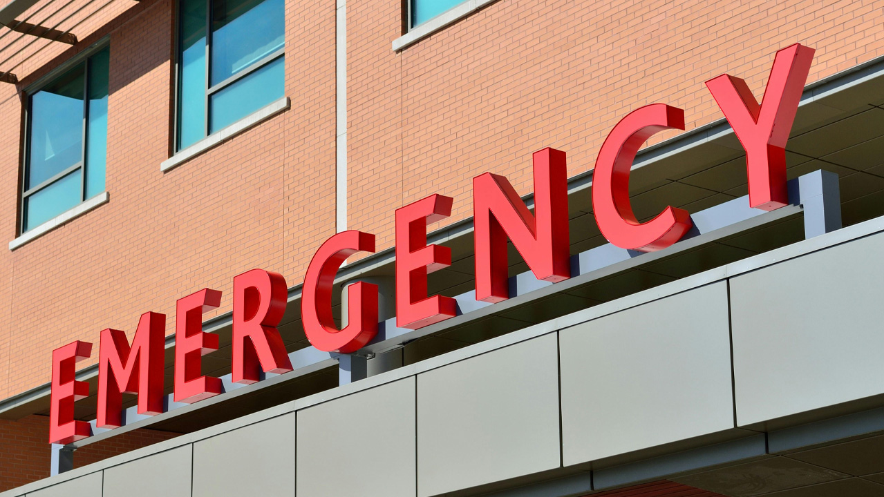 Photographie prise de jour d'une enseigne d’un service d’urgences hospitalières avec le mot « EMERGENCY » en grandes lettres rouges en relief, fixées sur la façade d’un bâtiment moderne en briques claires.